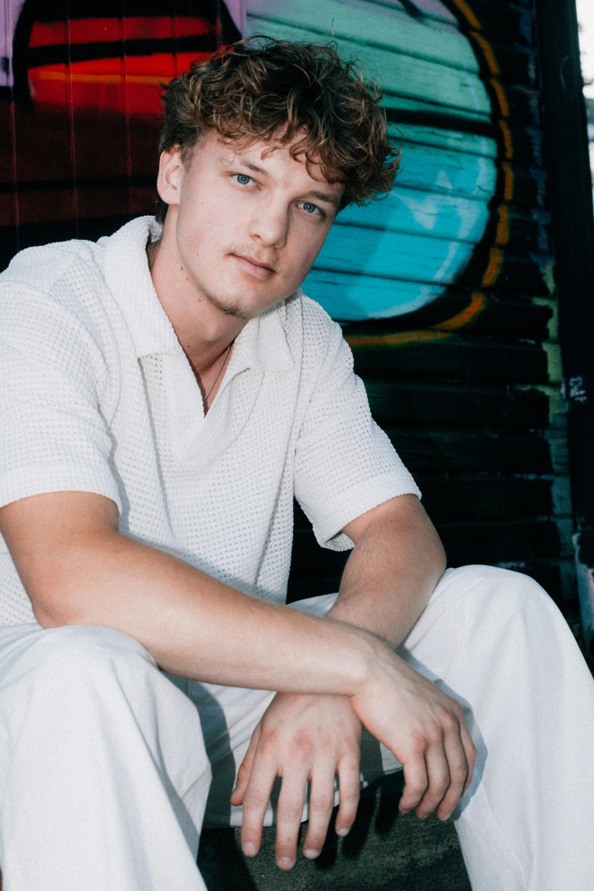 Man in a white outfit sitting against a colorful mural.